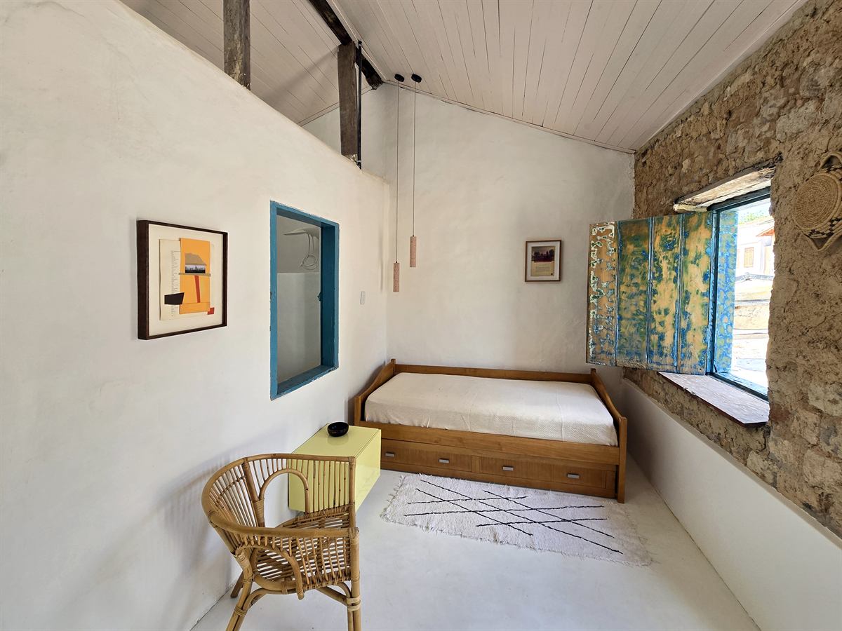 Quarto da Casa Tempo Glória com parede de pedra, janela azul, cama baixa de madeira e poltrona de vime
