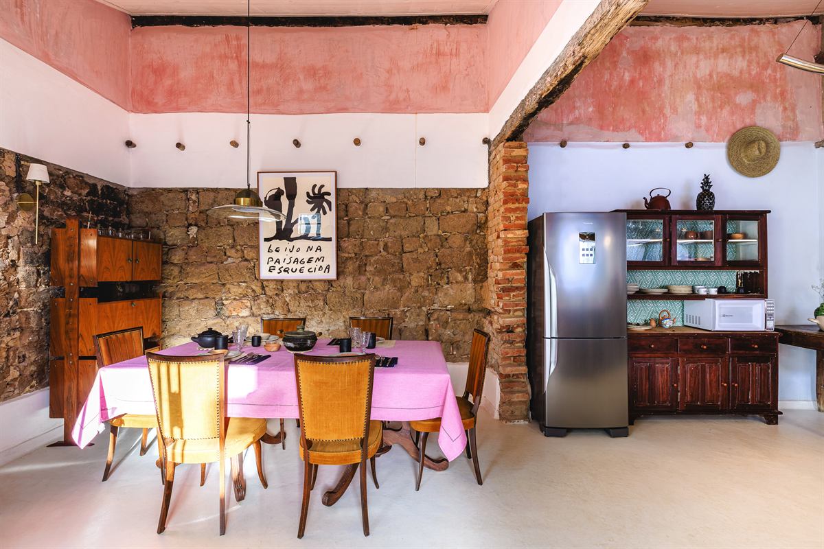 Mesa posta com toalha rosa e cadeiras de madeira, em cozinha com parede de pedra e cristaleira antiga