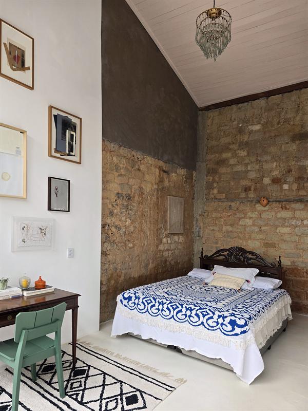 Quarto com cama de colcha azul e branca, parede de tijolos, quadros e cadeira verde