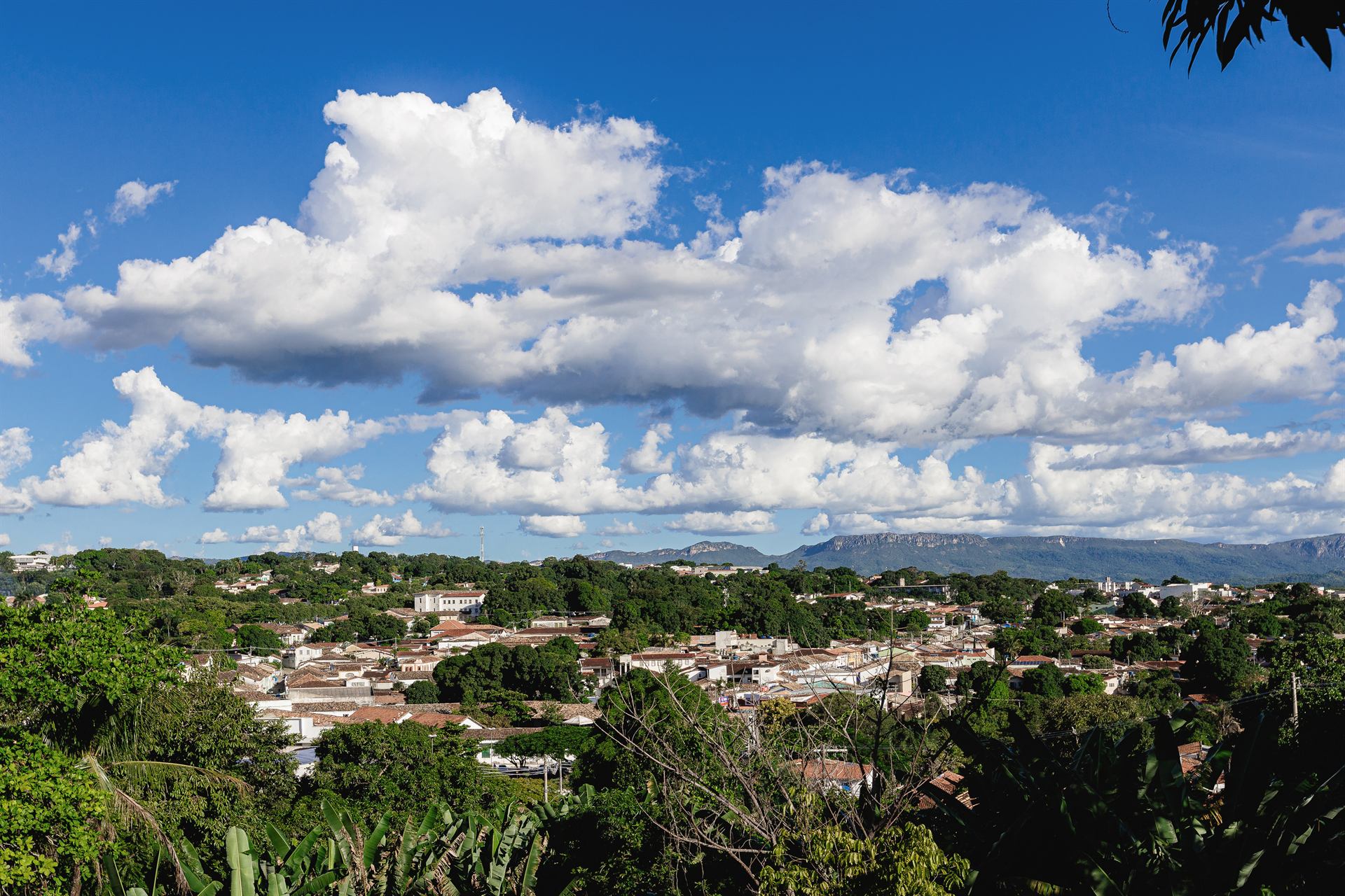 Vista do alto da Cidade de Goiás, com telhados coloniais entre a mata e a serra sob um céu azul com nuvens