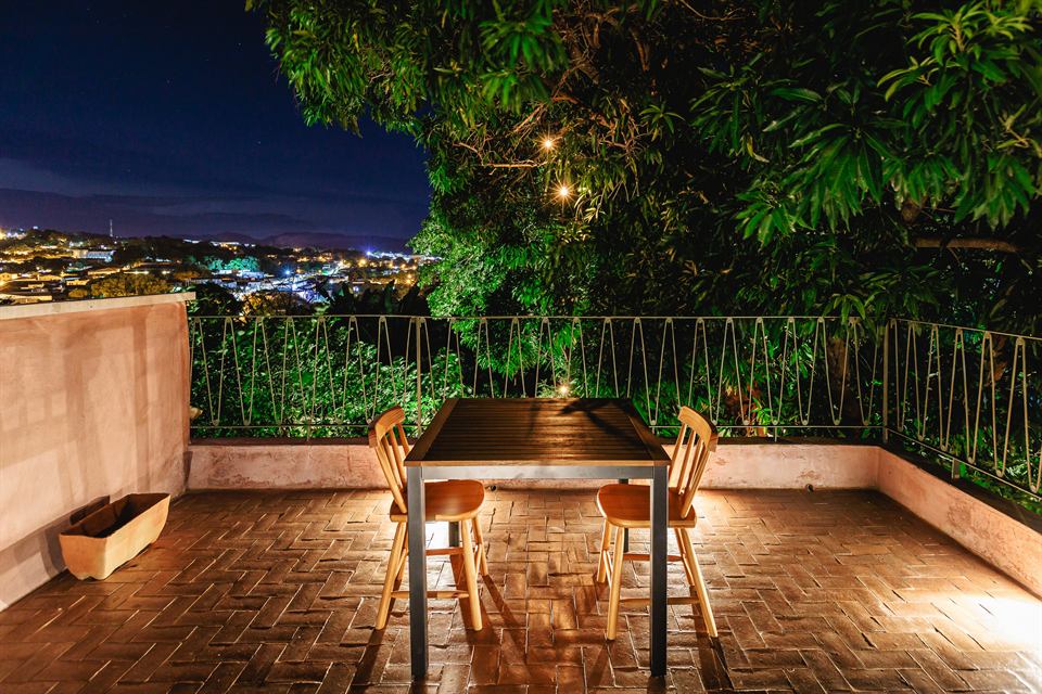 Varanda de piso de tijolos com mesa e cadeiras à noite, cercada de árvores, com as luzes da cidade ao fundo